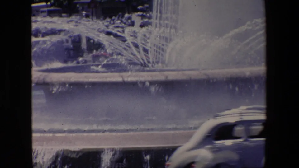 1967 Madrid home-movie frame showing a public fountain spraying water toward a road while a rounded car passes along the wet street.