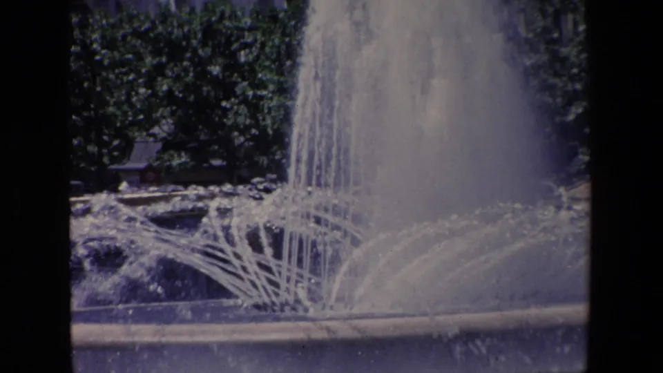 1967 Madrid frame with a large public fountain plume and lower arcing jets against trees, with the roadside edge visible in the foreground.