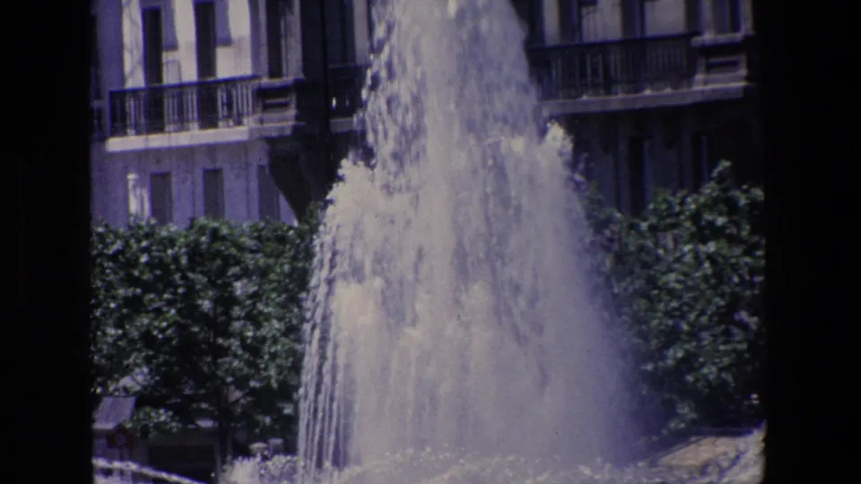 1967 Madrid 8mm frame showing a fountain plume with leafy trees and multi-story residential balconies in the background.