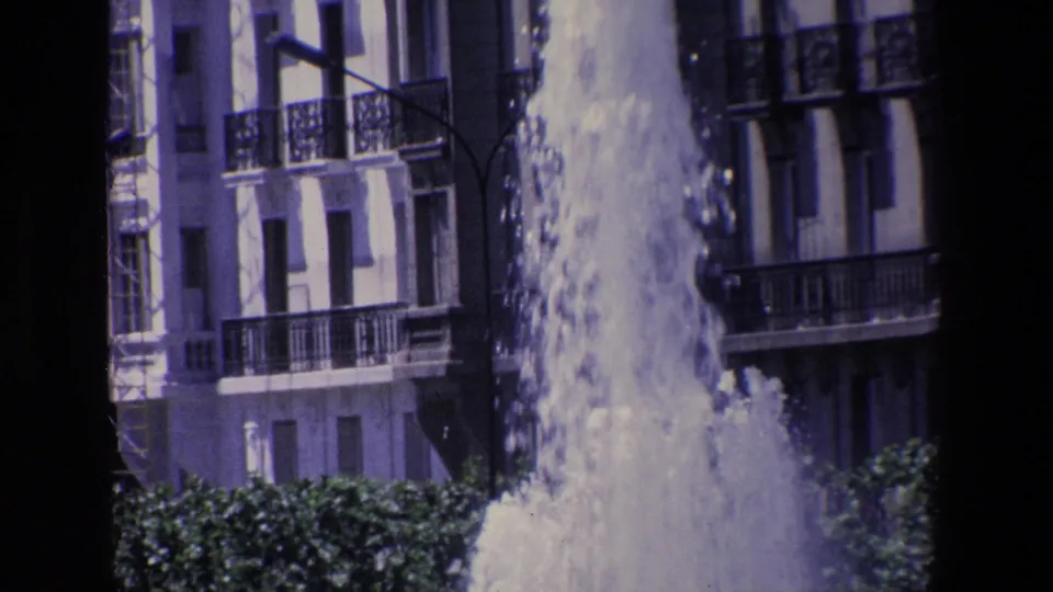 1967 Madrid frame where heavy fountain spray partially obscures trees and nearby apartment balconies in an urban setting.