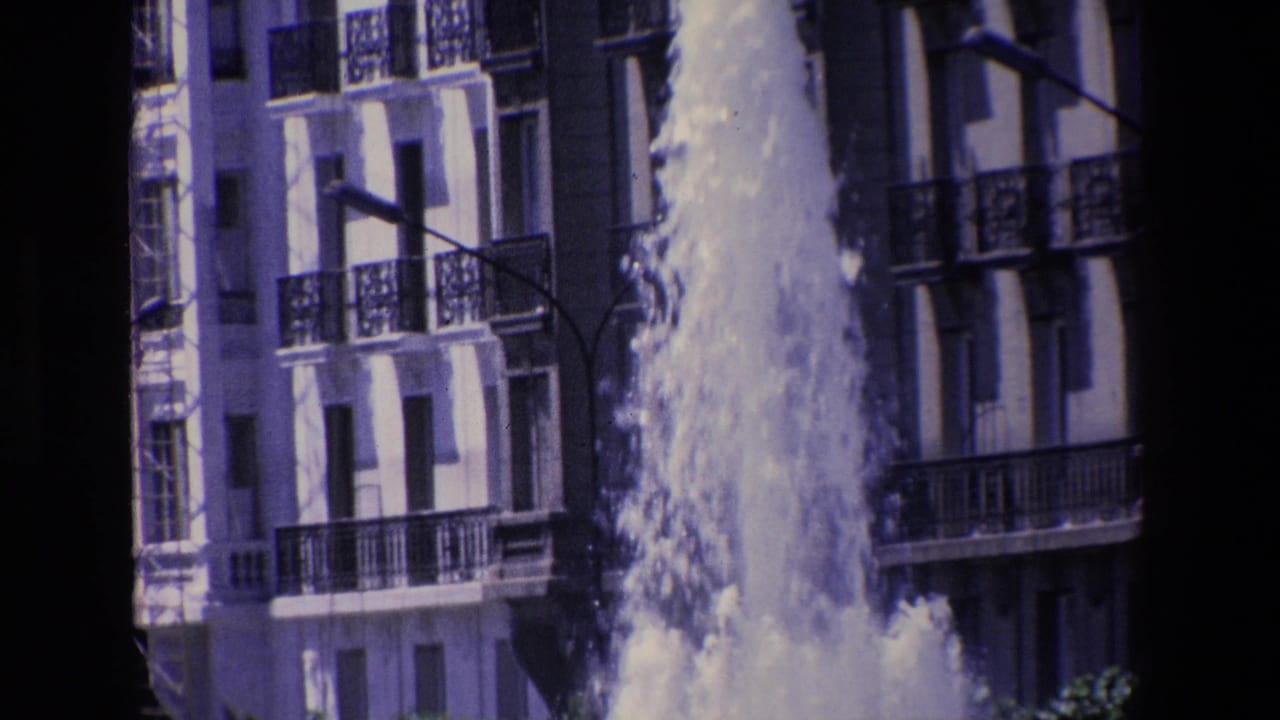 Water fountain rising in front of a multi-story building with balconies and a street light.
