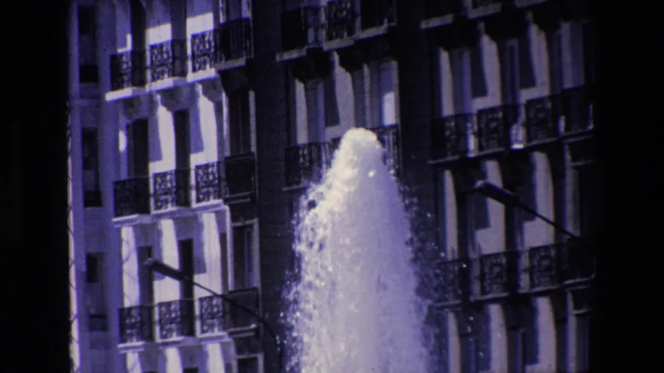 A lower portion of the fountain plume fills the foreground while the building facade, balcony railings, windows, and street light remain visible in shadowed contrast.