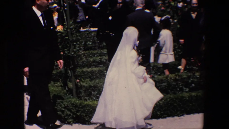 1967 Madrid wedding frame showing a bride in a white gown and veil walking past guests in dark formalwear near garden plantings.