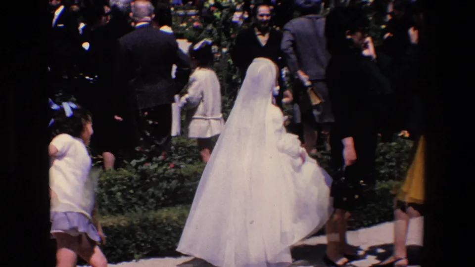 In 1967 Madrid home-movie footage, a young girl in a white communion dress and veil moves through a sunlit garden among adults and children.