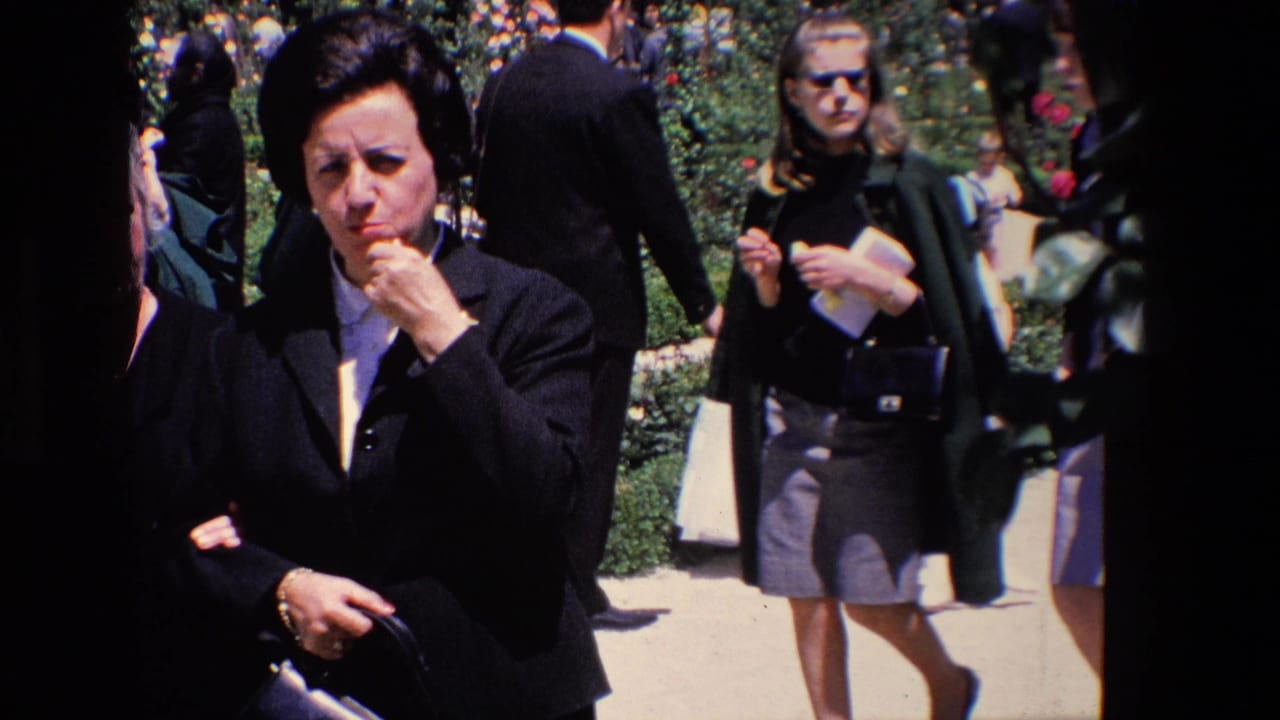 A woman in dark formal clothing stands close to the camera in a Madrid garden scene while other adults and children move behind her during the communion outing.