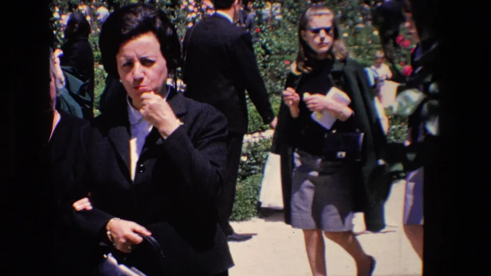 A woman in dark formal clothing stands close to the camera in a Madrid garden scene while other adults and children move behind her during the communion outing.