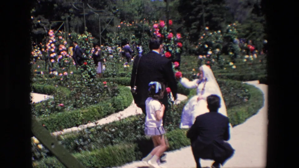 The young communion girl in a white dress appears beside rose beds in a Madrid garden as suited adults and children stand or crouch nearby.