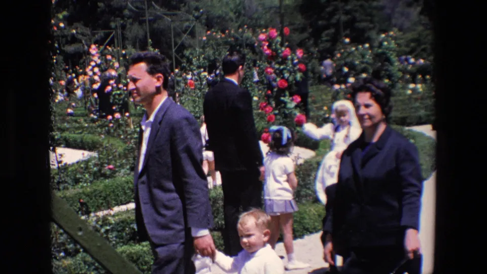 Adults and a small child cross the foreground of a 1967 Madrid garden scene while the communion girl in white remains visible among roses and family members behind them.