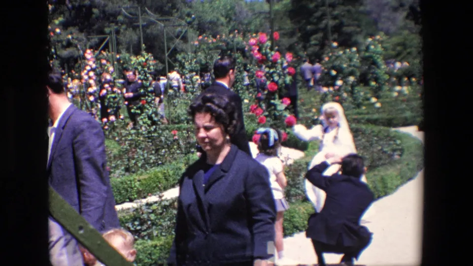 1967 Madrid home-movie frame showing a bride in a white gown near a garden path while guests and a crouching photographer gather among flower beds.