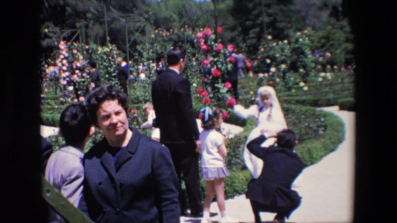 1967 Madrid wedding footage frame with a woman looking toward the camera, a bride posed farther back, and guests standing beside colorful flower beds.