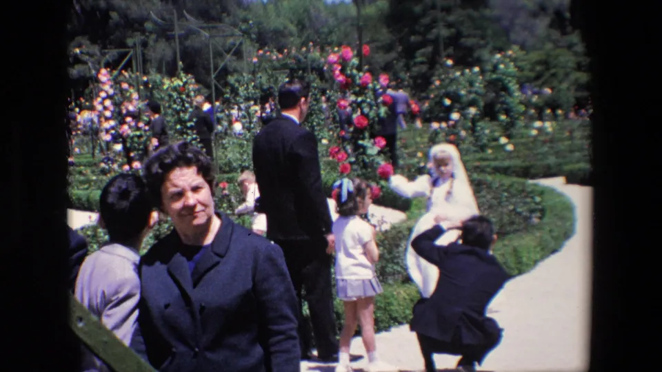 1967 Madrid wedding footage frame with a woman looking toward the camera, a bride posed farther back, and guests standing beside colorful flower beds.
