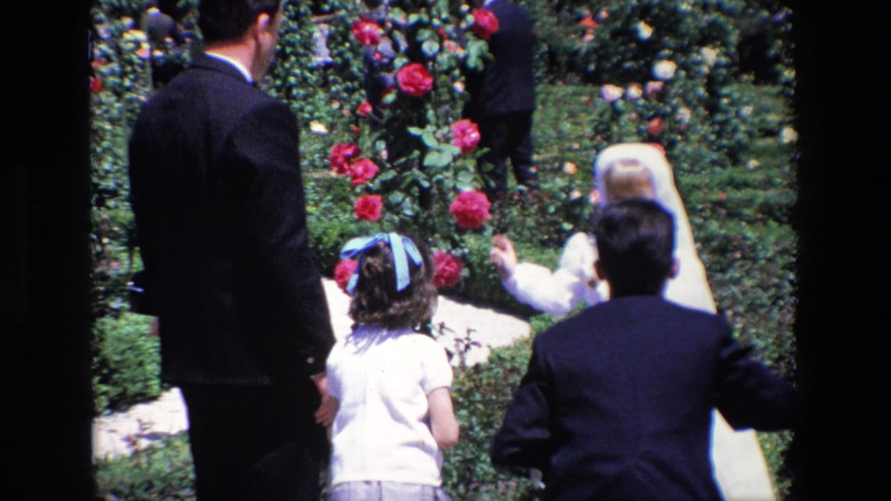In 1967 Madrid home-movie footage, adults in dark suits and children in dress clothes move along a garden path lined with roses.