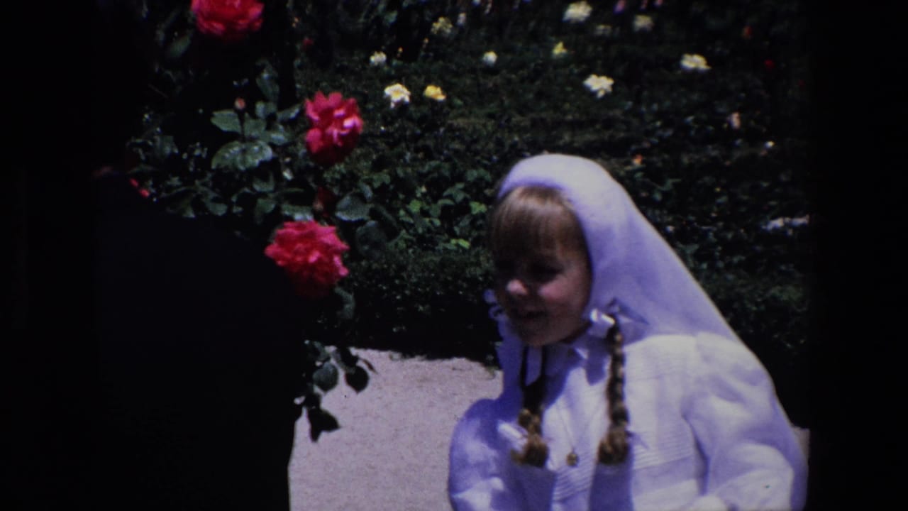 Young girl in a white veil stands near red roses and a garden path.
