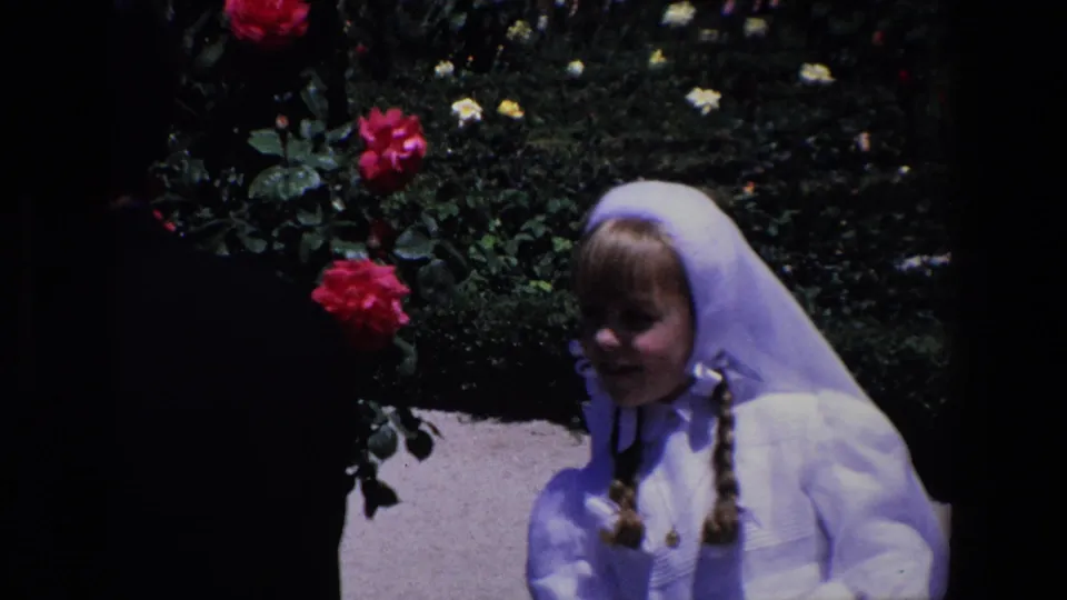 A young girl in a white dress and veil stands near rose bushes and a paved garden path in the Madrid home-movie scene.
