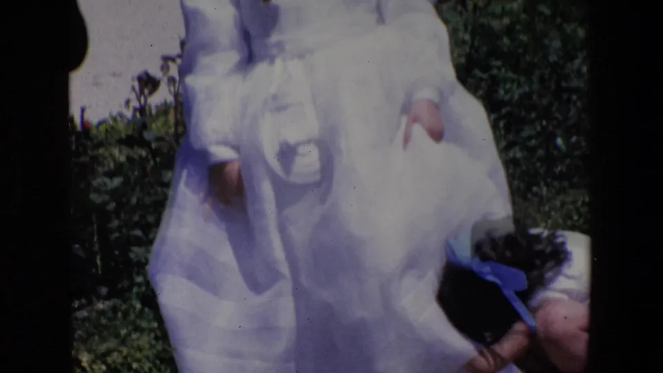 A close, slightly cropped home-movie frame shows the white dress and veil of a child, with another small child partly visible near the garden edge.
