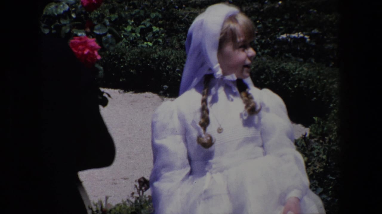 Madrid 1967 home-movie frame of a young girl in a white dress and veil-like head covering seated near rose bushes and a paved path.
