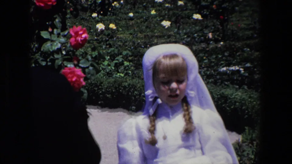 Madrid 1967 8mm frame showing a young girl in white formal clothing facing the camera, with braided hair, a head covering, roses, and garden planting behind her.
