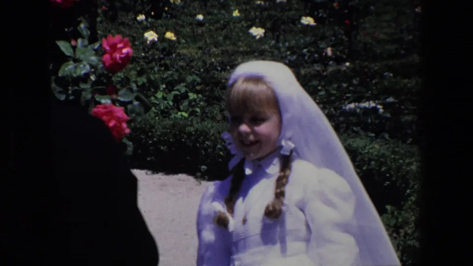 Madrid 1967 home-movie frame of a smiling young girl in a white dress and head covering seated or leaning near rose bushes in a sunny garden.
