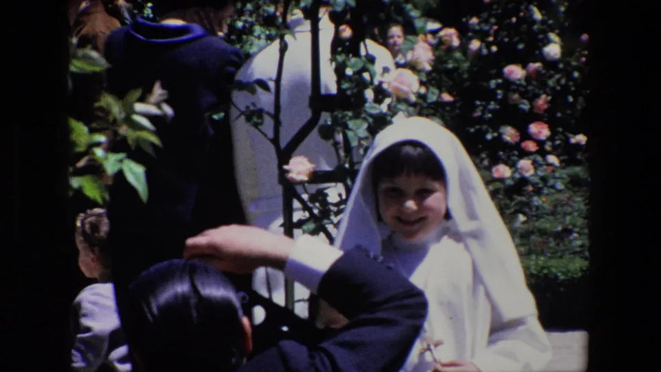 A young girl in white ceremonial clothing smiles toward the camera area as an adult in a dark suit raises a camera or hand in a sunlit Madrid garden.