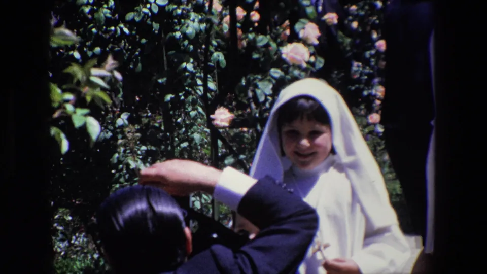 The frame shows a smiling young girl in a white veil and dress standing among rose bushes while an adult in a dark suit appears to compose a photograph.