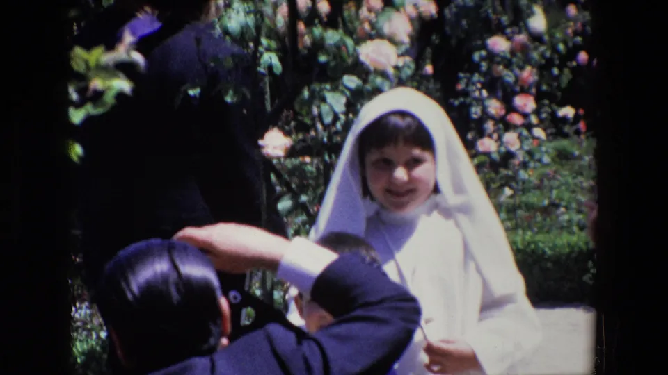 A smiling young girl in a white veil and formal dress stands in a rose garden in Madrid, with adults partly visible in the foreground and background.