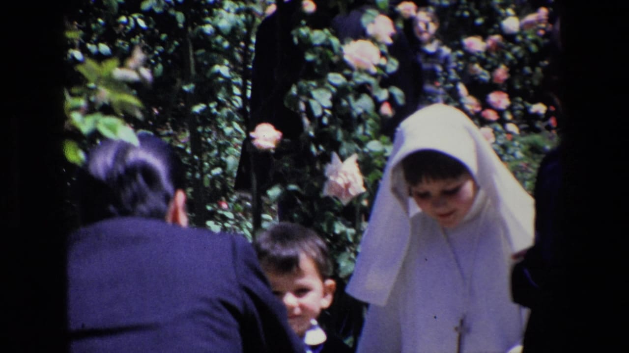 1967 Madrid home-movie frame showing children and adults close to rose bushes, with one child wearing a white veil and cross pendant.