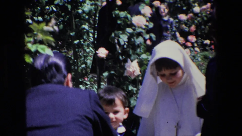 1967 Madrid home-movie frame showing children and adults close to rose bushes, with one child wearing a white veil and cross pendant.