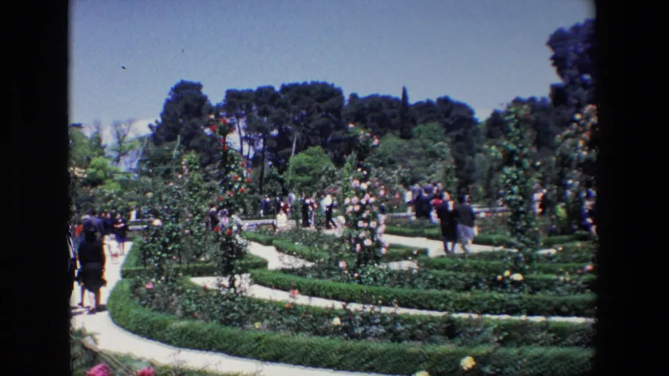 1967 Madrid frame showing visitors dispersed through a formal rose garden, with flowering shrubs, clipped hedges, and tree-lined background.