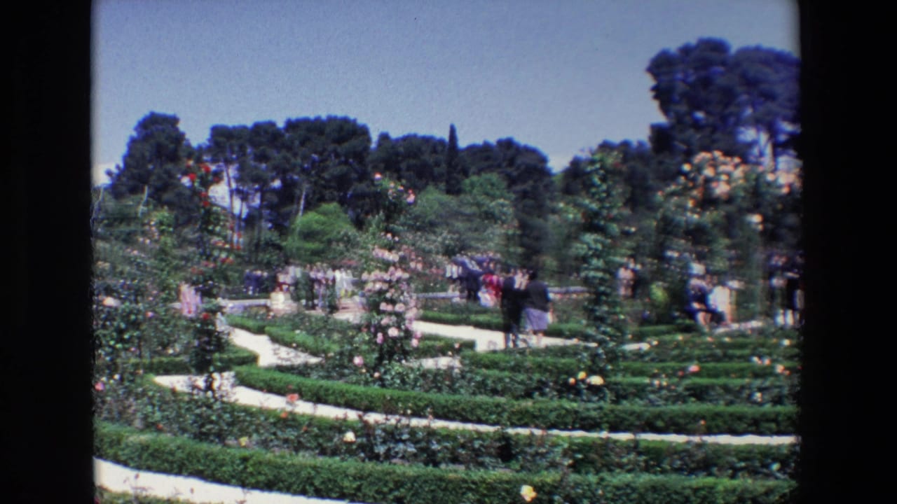 Visitors walking through a formal garden with hedges, flower beds, trees, and a bright sky.