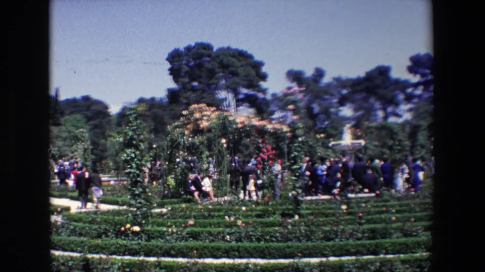 1967 Madrid home-movie frame showing people gathered along a garden path near rose-covered arches, clipped hedges, flowering shrubs, and tall trees.