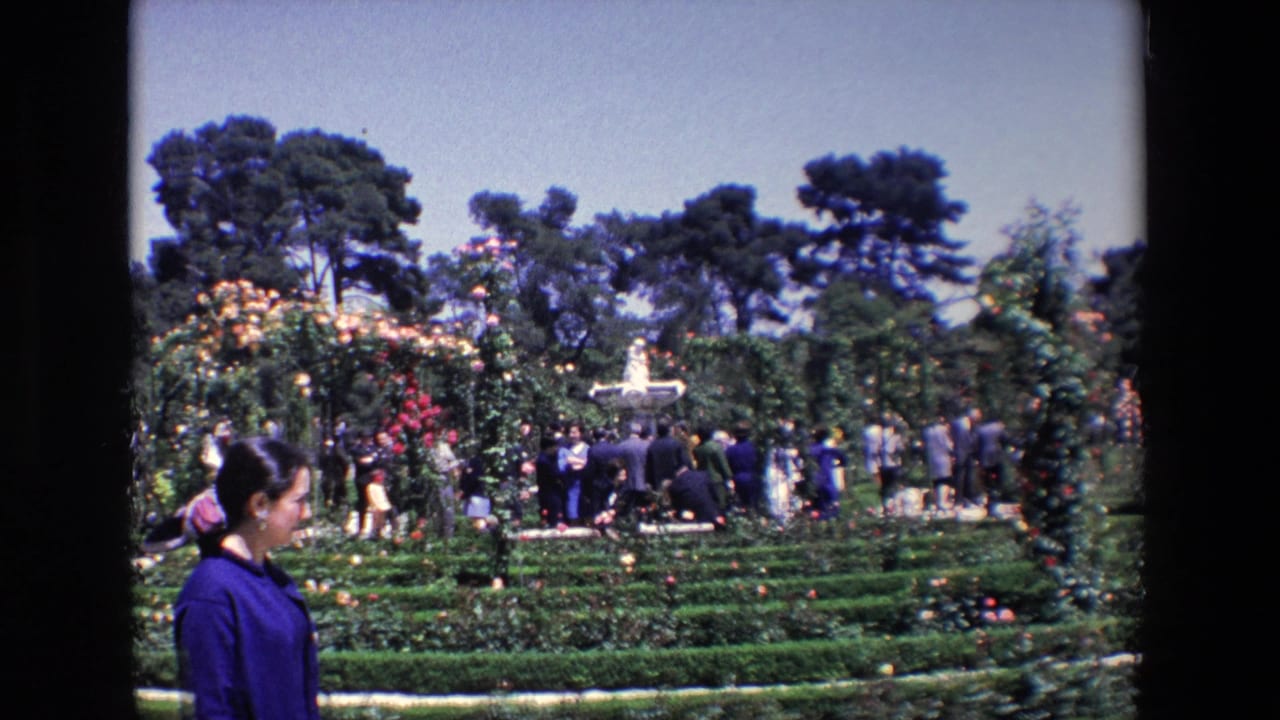 A woman in blue passes close to the camera as visitors stand among hedges, flowers, trees, and a fountain in the 1967 Madrid garden scene.