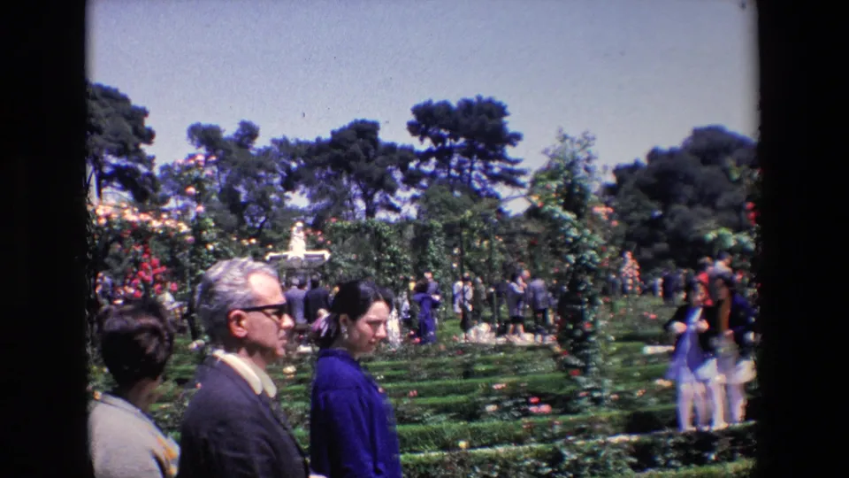 Foreground adults in period clothing move past the camera, with a landscaped Madrid garden, fountain, hedges, and other visitors visible behind them.