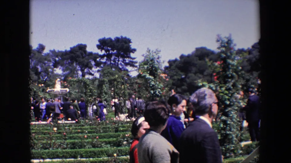 Adults and a possible child or young visitor stand near clipped hedges in the 1967 Madrid garden, with a fountain, trees, and other guests in the background.
