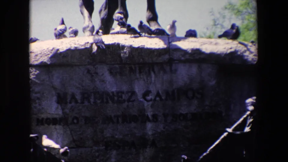 The frame moves closer to the monument base, where pigeons stand along the ledge above the partly shadowed Martinez Campos inscription.