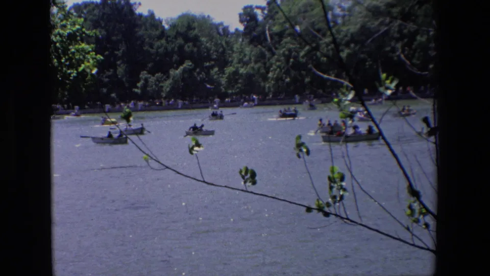 Wide home-movie view of a Madrid park lake, showing multiple rowboats with distant occupants against a tree-lined shore and overhanging branches.