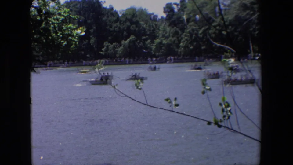 End frame of the Madrid park lake, with small boats moving across rippled water beneath dense trees and thin leafy branches in front of the lens.