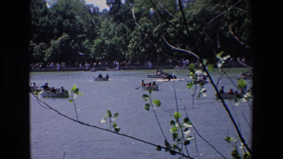 1967 Madrid home-movie still showing multiple small boats with people on open water, partly obscured by foreground branches and leaves.