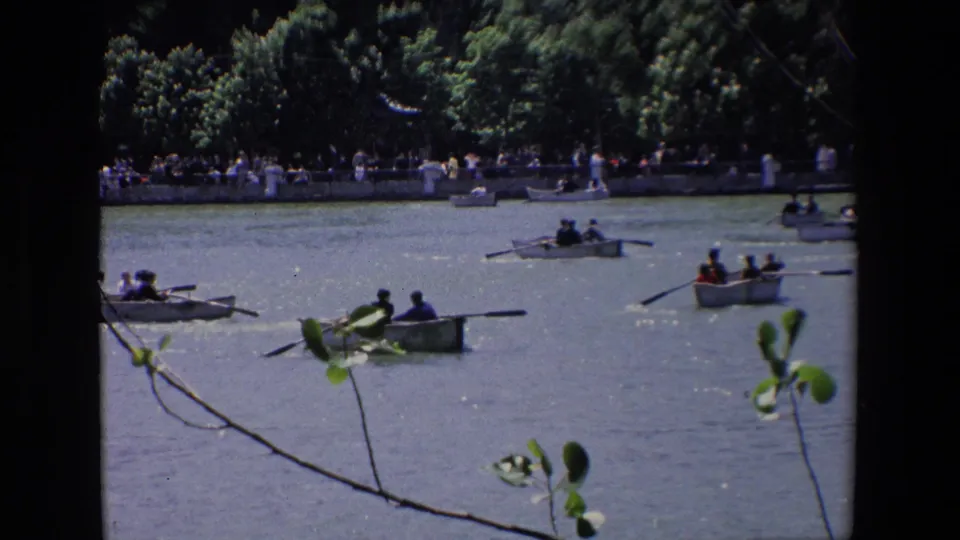 1967 Madrid home-movie frame showing several small rowboats with passengers on sunlit water, seen through foreground leaves with people gathered along the far bank.