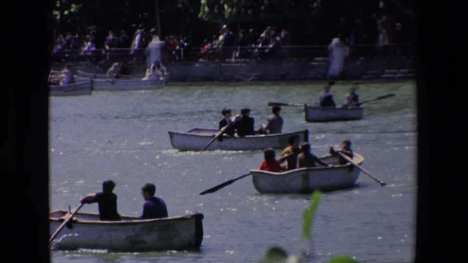 Home-movie view of several rowboats carrying adults and possible children, with a crowded bank and trees behind them in the Madrid leisure setting.