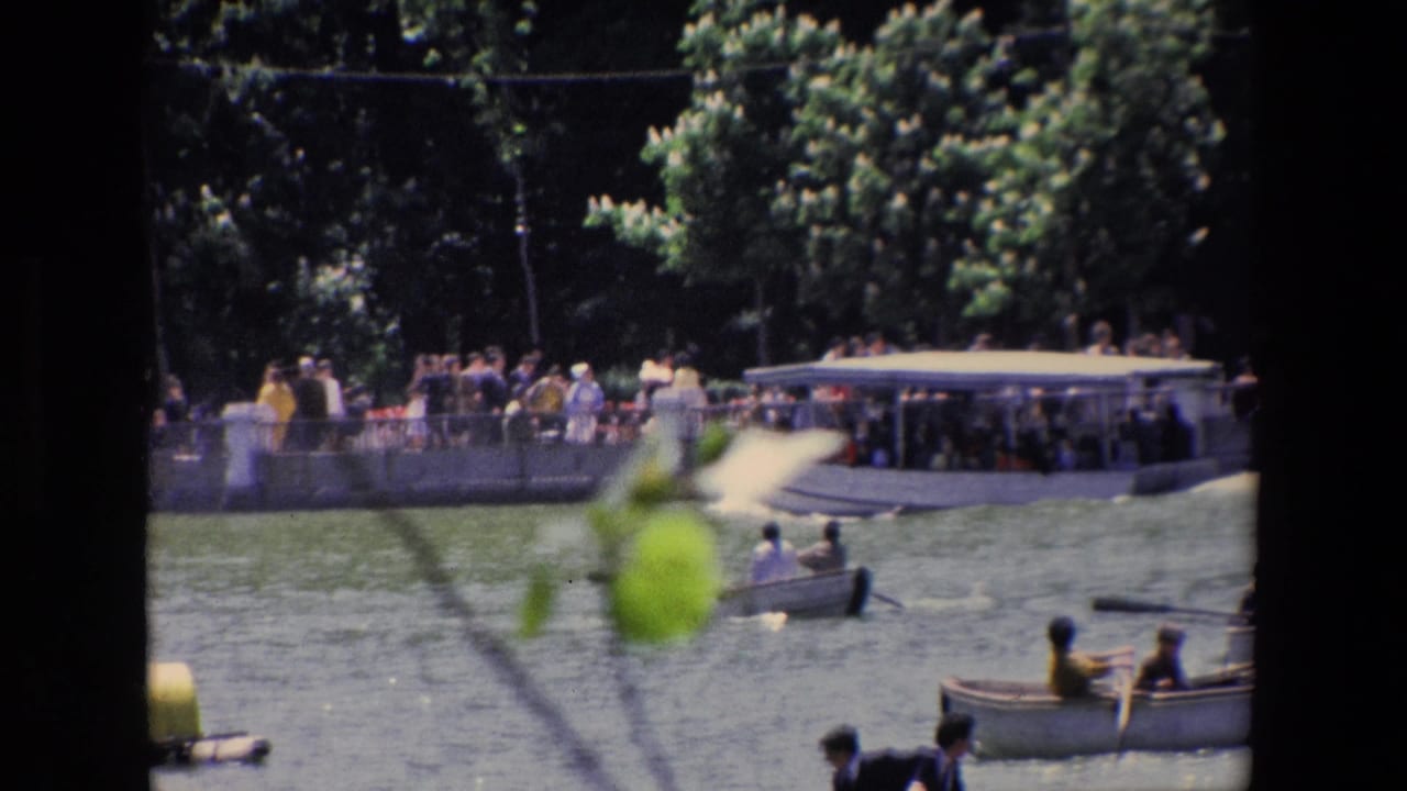1967 Madrid home-movie frame showing visitors beside a lake with small rowboats, dense trees, and a waterside structure.