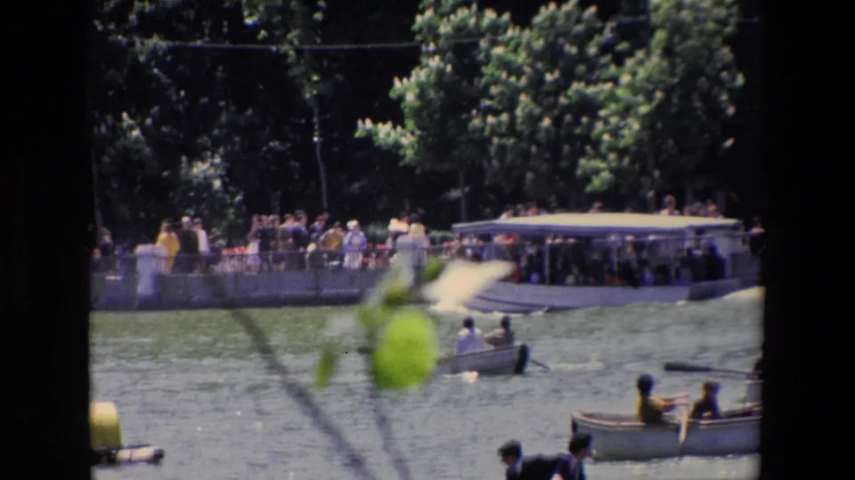 1967 Madrid home-movie frame showing visitors beside a lake with small rowboats, dense trees, and a waterside structure.