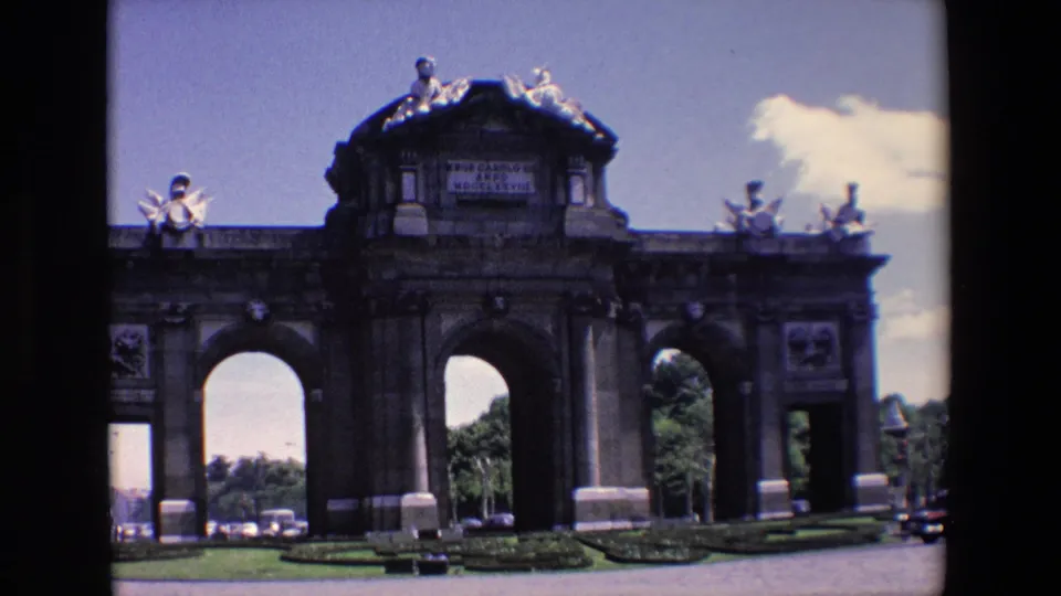 1967 Madrid frame centered on the Puerta de Alcala, showing its large arches, upper inscription area, stone ornament, roadway, and surrounding greenery.
