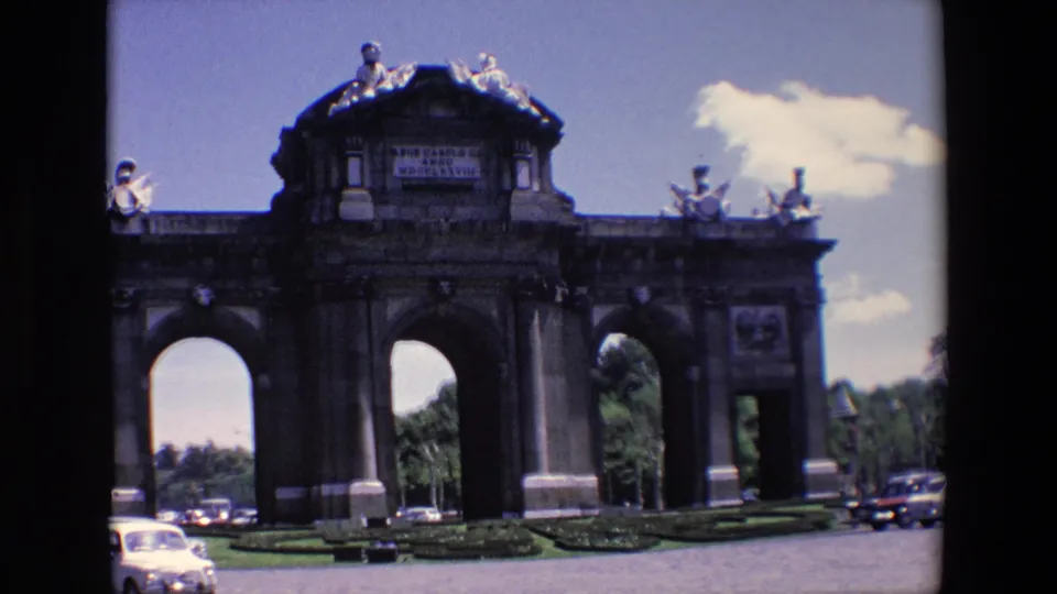 1967 Madrid home-movie frame of the Puerta de Alcala, with classic cars along the road, low lawns, stone arches, and bright sky.