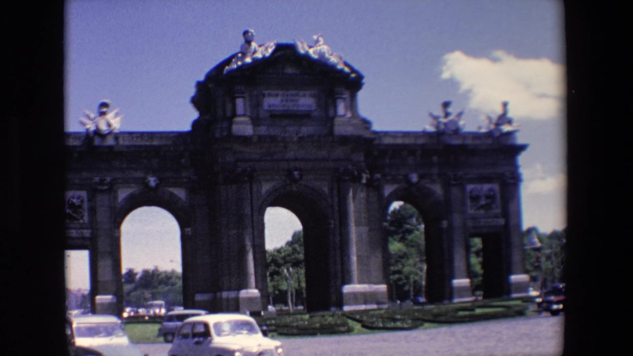 Stone archway with rooftop statues and cars passing in front under a blue sky.