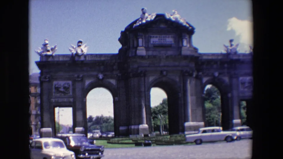 1967 Madrid home-movie view of Puerta de Alcala with the camera closer to the facade as mid-century cars pass below the arches.