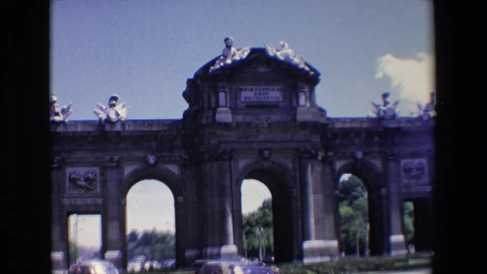 1967 Madrid frame showing Puerta de Alcala from across the roadway, with cars at the base and the monument's sculptural roofline silhouetted against the sky.