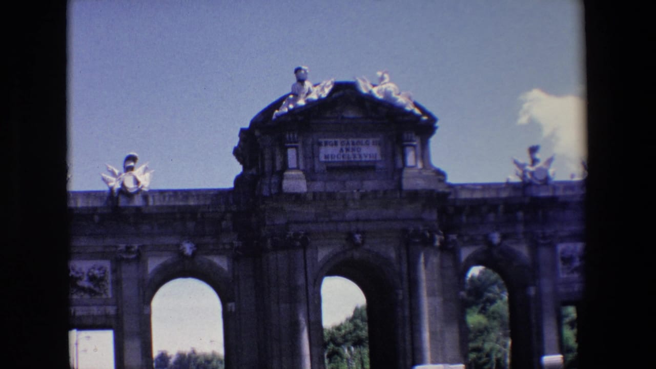Wide view of a stone arched gate with rooftop statues and a Latin inscription under blue sky.