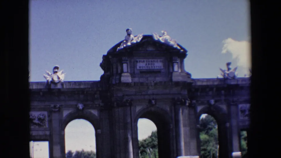 1967 Madrid home-movie frame showing Puerta de Alcala from below, with arched openings, sculptural roof figures, and a visible dedication panel.