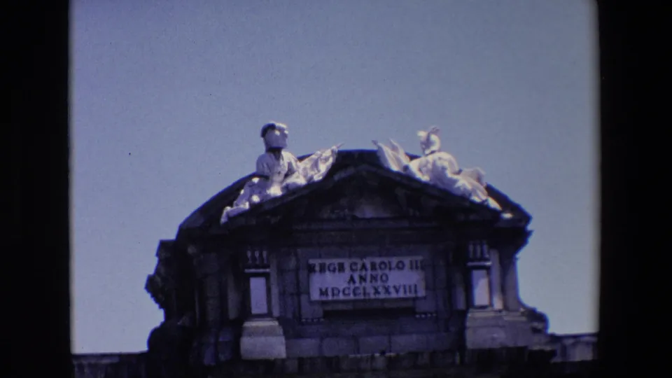1967 Madrid home-movie frame showing a closer look at Puerta de Alcala's sculpted roofline, inscription panel, and heavy stonework.