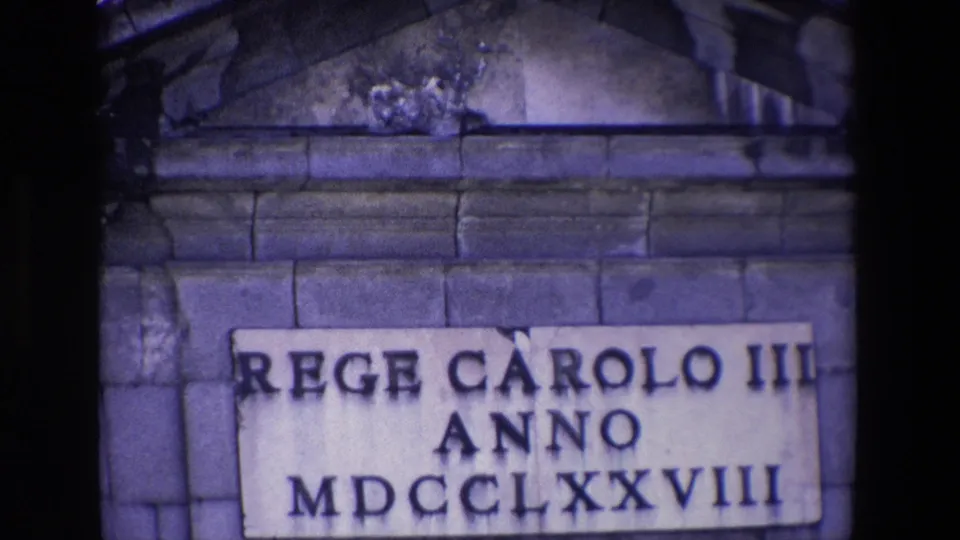 Madrid 1967 home-movie frame with a closer view of a weathered plaque reading REGE CAROLO III ANNO MDCCLXXVIII, set into block stonework beneath classical architectural trim.